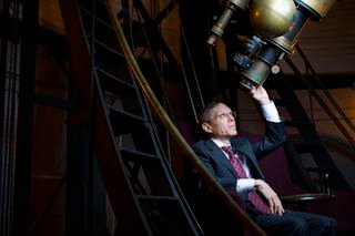 A man in a suit sitting on the floor looks through a large telescope